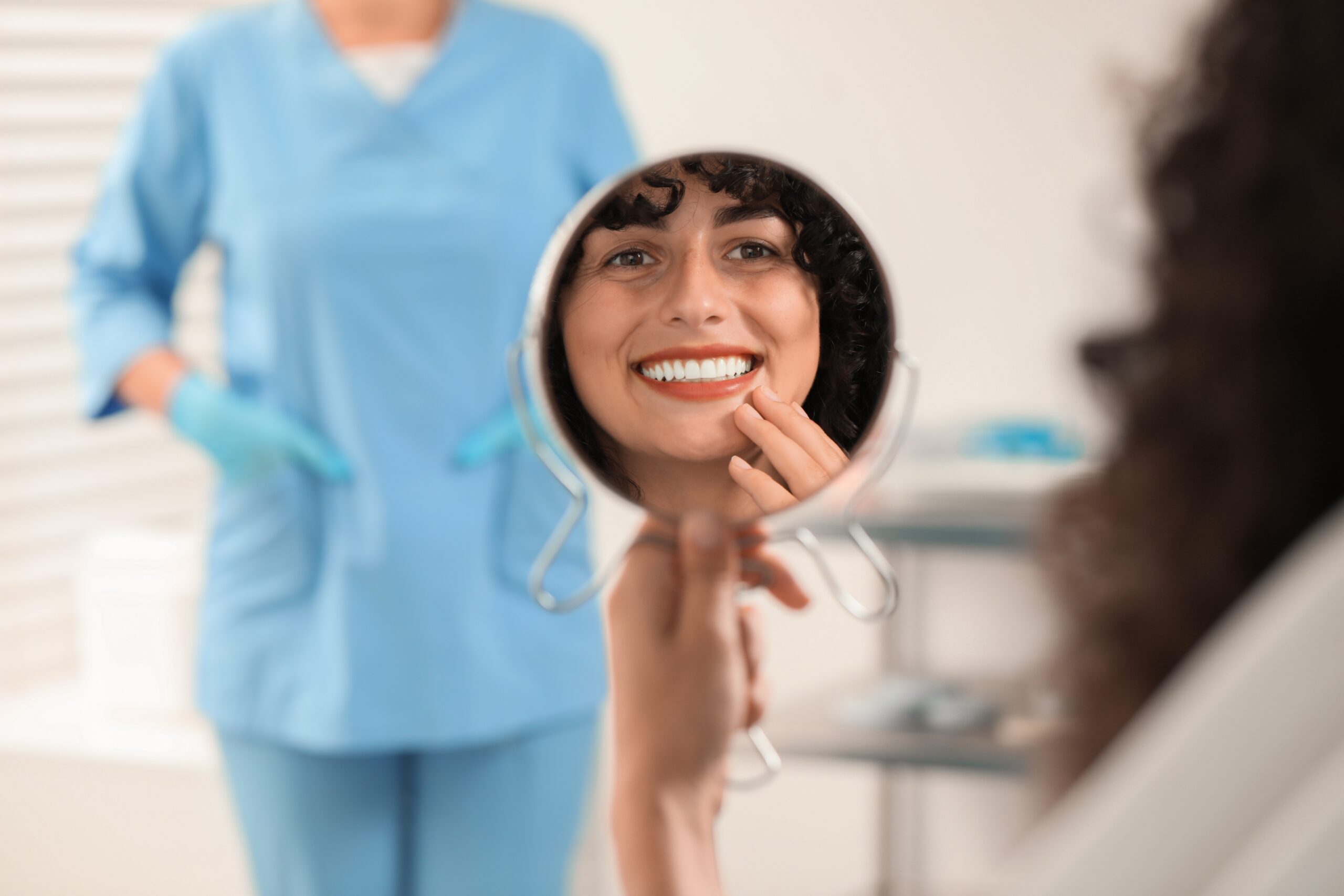 woman smiling in a small mirror with dentist in background, happy with new veneers